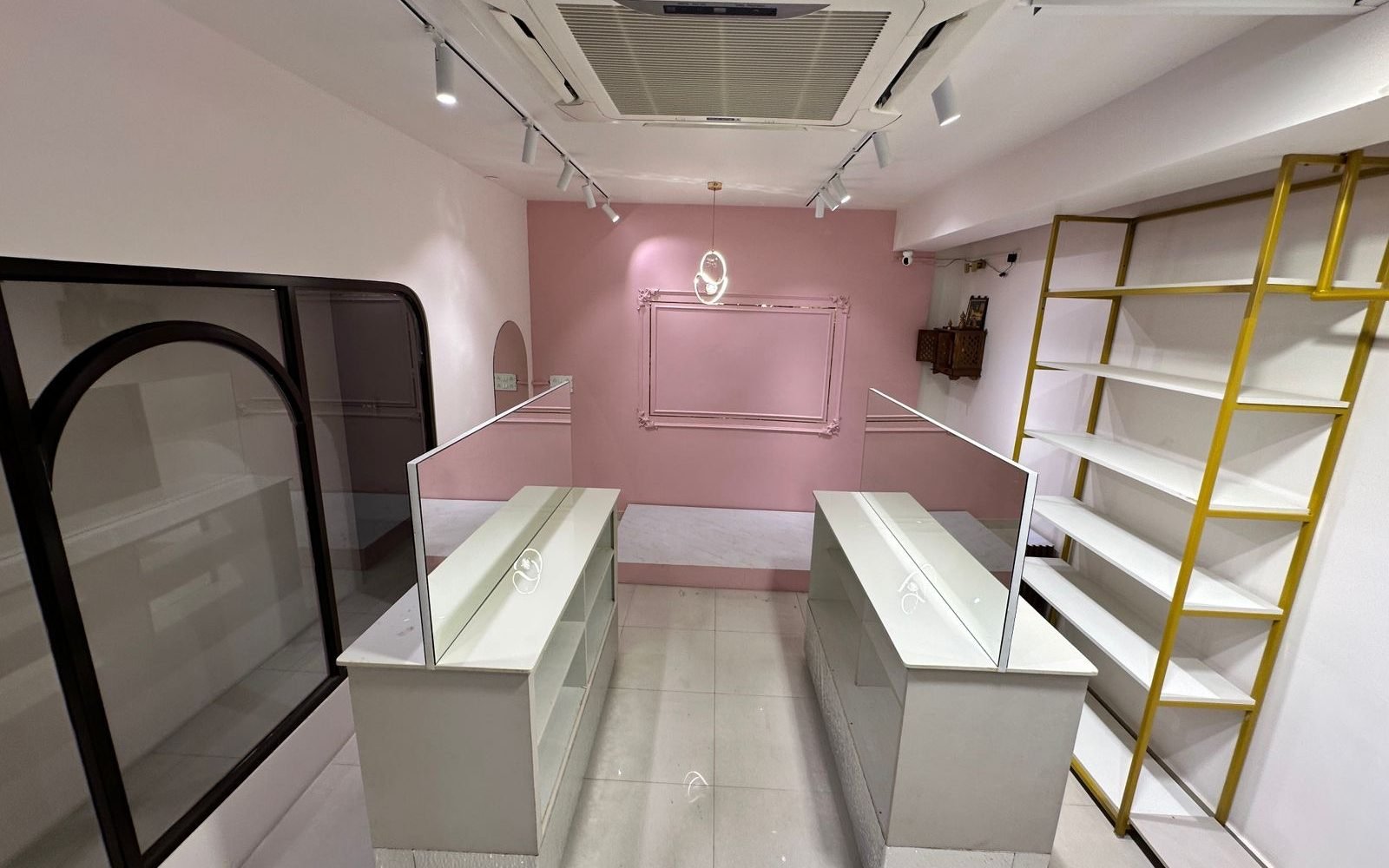 The Ananta Designs boutique in Bangalore seen from the entrance: a display counter on each side, gold shelving along the right wall, and the pink feature wall at the far end.