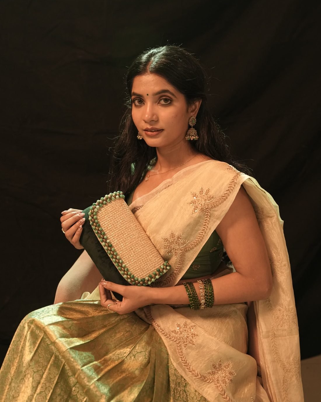 A model in a cream and gold saree with a green blouse and green bangles, against a dark backdrop.