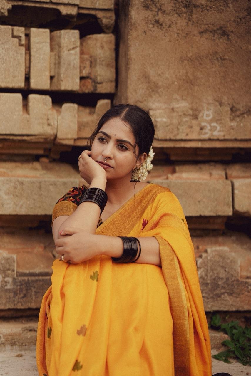 A model seated in a mustard yellow saree with a printed blouse, black bangles and jasmine in her hair, before carved stone ruins.